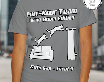 May include: Grey t-shirt featuring the text "Purr-Kour Team Living Room Edition" and a graphic of cats leaping over a sofa and curtain. The words "Sofa Gap - Level 7" are also visible.