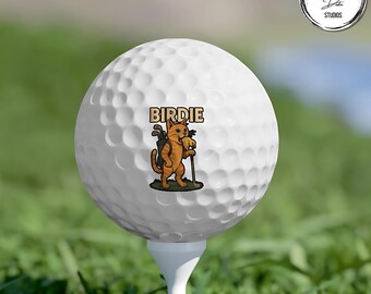 May include: White golf ball on a tee with a cartoon cat graphic. The cat is orange and brown, holding golf clubs and a golf ball, with the word "Birdie" above it. The ball is on a green golf course.