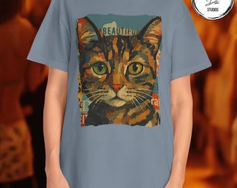 May include: A light blue t-shirt featuring a colorful, artistic portrait of a cat's face. The cat has green eyes and a brown and orange coat. The word "BEAUTIFUL" is visible above the cat's head.