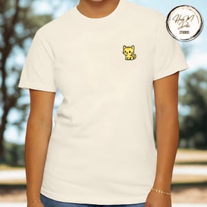 May include: Off-white t-shirt with a small embroidered cartoon cat in yellow and black on the left chest. The shirt has short sleeves and a crew neck. A circular logo with text is visible on the upper right.