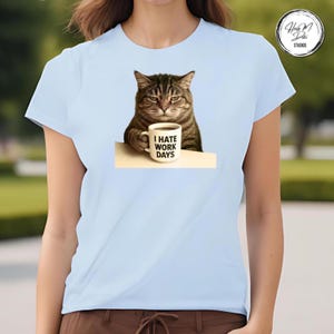 May include: Light blue t-shirt featuring a grumpy cat holding a coffee mug. The mug has the text "I HATE WORK DAYS" printed on it. The cat is brown and gray with a displeased expression.