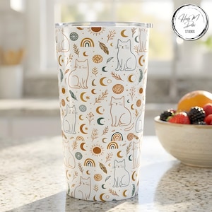 May include: White tumbler with a repeating pattern of cats, moons, rainbows, and suns in brown and teal. The tumbler has a silver lid and is on a kitchen countertop. The design is whimsical and bohemian.
