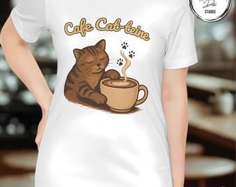 May include: White t-shirt featuring a brown tabby cat enjoying a cup of coffee. The phrase "Cafe Cat-feine" is written above the cat in a whimsical font. The design includes paw prints and steam rising from the coffee cup.