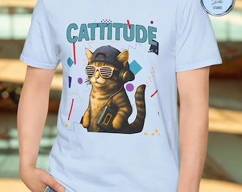 May include: Light blue t-shirt featuring a cartoon cat wearing sunglasses, a cap, and headphones, with a boombox. The word "Cattitude" is printed above the cat in teal and blue. The shirt has a retro 80s and 90s vibe.