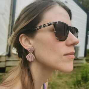 Può includere: Un paio di orecchini a forma di conchiglia rosa chiaro. Gli orecchini sono realizzati in un materiale liscio e hanno un design a strati. La persona indossa occhiali da sole con montatura maculata bianca e nera.