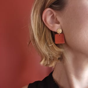 Peut inclure: Gros plan sur une oreille portant des boucles d'oreilles. Chaque boucle d'oreille est composée d'un petit disque doré texturé surmontant un pendentif carré de couleur terre cuite. Le fond est rouge chaud uni.