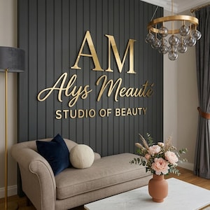 May include: A beige chaise lounge with a navy blue velvet pillow and a round cream textured pillow sits before a dark gray vertical slat wall featuring gold 3D lettering that reads AM Alys Meauté STUDIO OF BEAUTY, with a gold chandelier above.