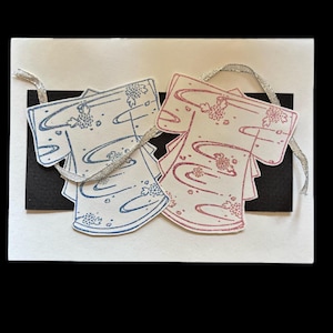 May include: Two paper kimono-shaped cards, one blue and one pink, are attached to a white card with a black border. The kimono cards have floral and wave patterns. Silver ribbon is attached to the top of the kimono cards.