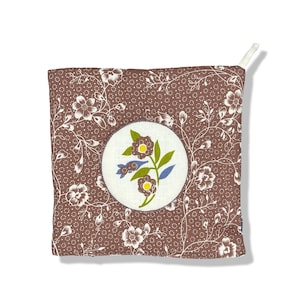 May include: Square brown pot holder with a white floral pattern and a central circular design. The circle features a floral illustration with green leaves, yellow centers, and blue accents. A white loop is attached to one corner.