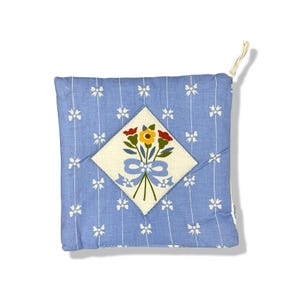 May include: A square, light blue pot holder with a floral design. The pot holder has a white diamond-shaped patch in the center with a bouquet of flowers in red, yellow, and green, tied with a blue ribbon. White bows and vertical stripes decorate the background.