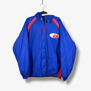 May include: A blue hooded jacket with red accents on the shoulders and cuffs. The jacket features a full-length zipper and a patch with a logo on the chest. The jacket is hanging on a black hanger against a white background.