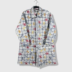 May include: A long-sleeved, button-down quilted coat featuring a white and navy blue plaid pattern. The fabric is decorated with a repeating print of various small, colorful cartoon creatures in shades of yellow, red, and blue. It has a collar.