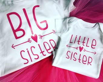 May include: Two white baby onesies with pink text that reads "Big Sister" and "Little Sister". The text includes arrows and hearts. The onesies are on a pink tulle skirt.