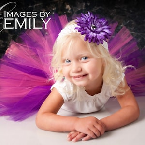 May include: A young girl with blonde hair wearing a white shirt and a pink and purple tutu. She is wearing a headband with a purple flower on it. The background is a dark gray.