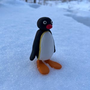 Puede incluir: Una pequeña figura de pingüino hecha a mano con arcilla, de pie en la nieve. El pingüino tiene un cuerpo negro, vientre blanco, ribete amarillo, pies naranjas y pico rojo. El fondo es un paisaje nevado con árboles.