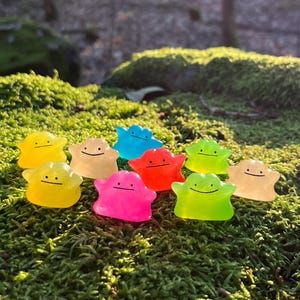 May include: A group of colourful, translucent Ditto figures in shades like yellow, blue, pink, and green. Each figure has a simple, smiling face. The figures are arranged on green moss, with a blurred forest backdrop.