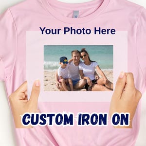 May include: A light pink t-shirt with the text "Your Photo Here" and "CUSTOM IRON ON". The shirt displays a family photo on the front. The photo shows a family of three on a beach.