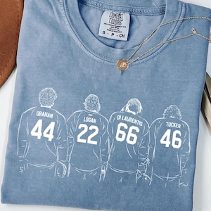 May include: A blue short-sleeved t-shirt featuring a white line-art graphic of four individuals wearing jerseys with the names Graham, Logan, Di Laurentis, and Tucker, along with the numbers 44, 22, 66, and 46. The shirt has a Comfort Colors brand label.