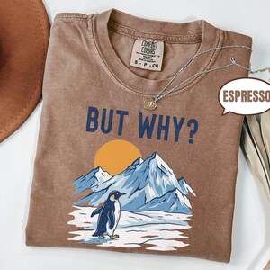 May include: Espresso-colored t-shirt with the phrase "BUT WHY?" in navy blue. The graphic features a penguin standing on ice in front of mountains and a yellow sun. A silver necklace is draped across the shirt.
