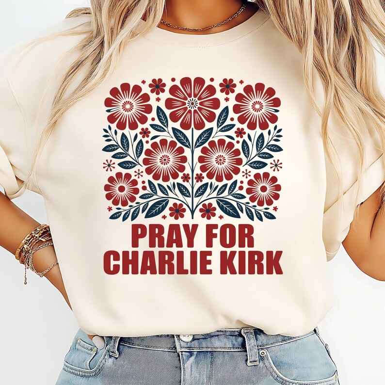 May include: Cream-colored t-shirt featuring a floral design in red and white, with dark blue leaves. The text "PRAY FOR CHARLIE KIRK" is printed in red block letters below the floral arrangement. The shirt has short sleeves.