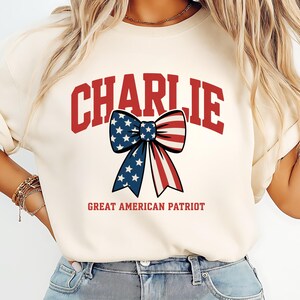 May include: Cream-colored sweatshirt with the name "CHARLIE" in red block letters. A patriotic bow graphic with blue stars and red and white stripes is centered below the name. The text "GREAT AMERICAN PATRIOT" is below the bow.
