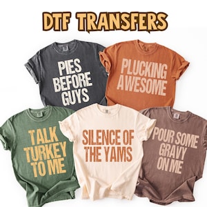 May include: A collection of five t-shirts in various colors, each with a unique Thanksgiving-themed phrase printed in a contrasting color. The phrases include "Pies Before Guys," "Plucking Awesome," and "Talk Turkey To Me."