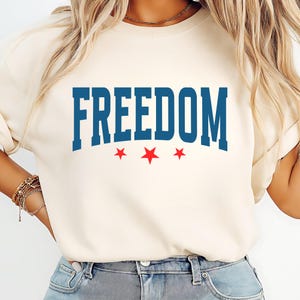 May include: Cream-colored t-shirt with the word "FREEDOM" in bold, blue, collegiate-style lettering. Below the word are three red stars. The shirt is a casual style.