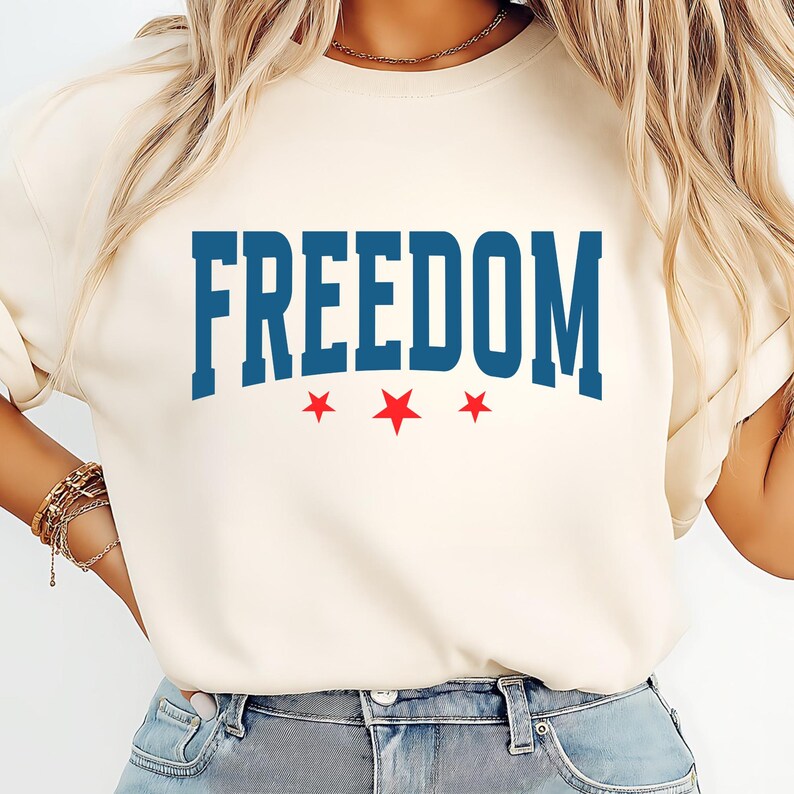 May include: Cream-colored t-shirt with the word "FREEDOM" in bold, blue, collegiate-style lettering. Below the word are three red stars. The shirt is a casual style.