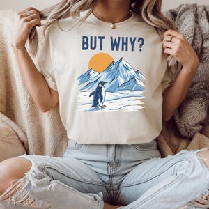 May include: Beige t-shirt with the text "BUT WHY?" above a graphic of a penguin standing on ice in front of mountains and a sun. The penguin is black and white, the mountains are blue, and the sun is orange.