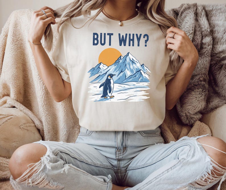 May include: Beige t-shirt with the text "BUT WHY?" above a graphic of a penguin standing on ice in front of mountains and a sun. The penguin is black and white, the mountains are blue, and the sun is orange.