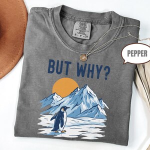 May include: Gray t-shirt with the text "BUT WHY?" above a graphic of a penguin, mountains, and a sun. The shirt has a crew neck and short sleeves. A gold necklace is draped across the shirt.