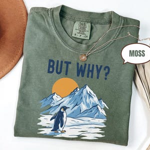 May include: A moss green t-shirt with the text "BUT WHY?" above a graphic of a penguin, mountains, and a setting sun. The shirt has a crew neck and short sleeves. A gold necklace is draped across the shirt.