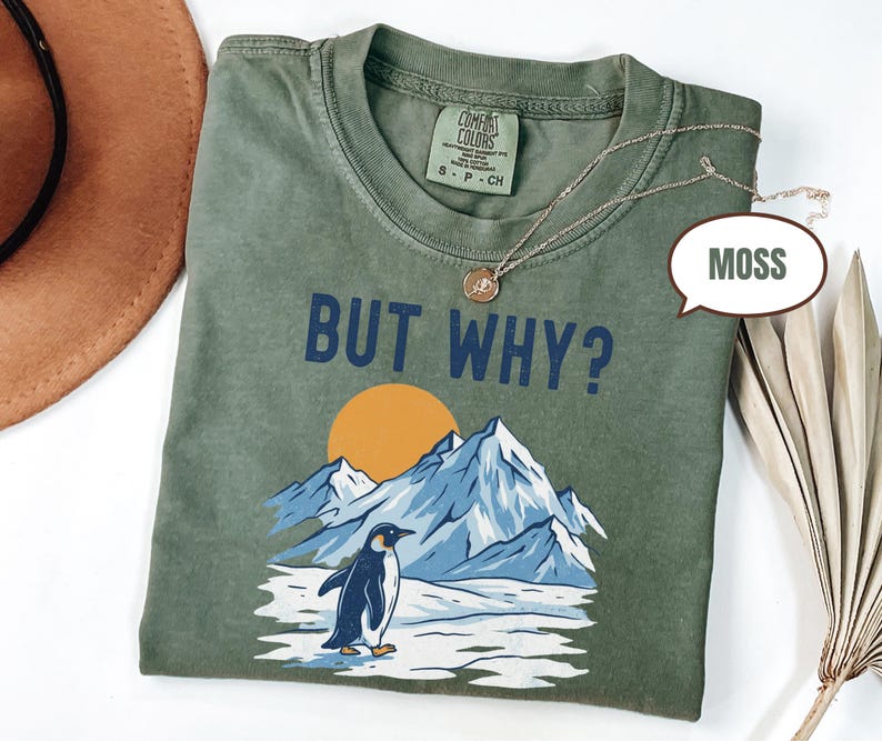 May include: A moss green t-shirt with the text "BUT WHY?" above a graphic of a penguin, mountains, and a setting sun. The shirt has a crew neck and short sleeves. A gold necklace is draped across the shirt.
