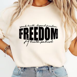 May include: Cream-colored t-shirt with the word "FREEDOM" in bold black letters. Above the word is "speak truth, depend freedom", and below is "A truth patriot". The shirt also has the text "1990-2025" and "Charlie Kirk".