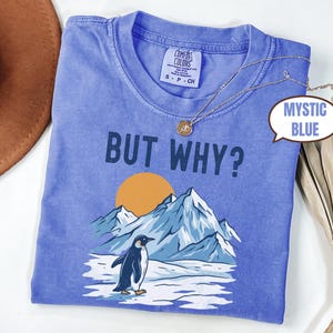 May include: Mystic blue t-shirt with the phrase "BUT WHY?" above a graphic of a penguin standing in front of mountains and a setting sun. A gold necklace is draped across the shirt.