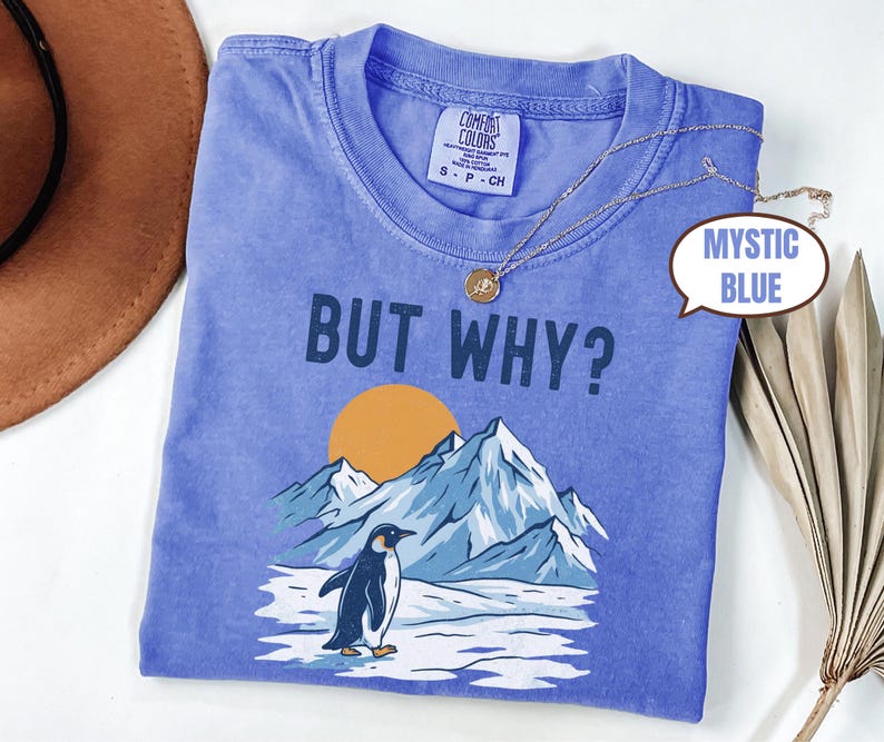 May include: Mystic blue t-shirt with the phrase "BUT WHY?" above a graphic of a penguin standing in front of mountains and a setting sun. A gold necklace is draped across the shirt.