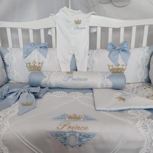 Peut inclure: Ensemble de literie pour bébé blanc et bleu clair, avec broderie de couronne dorée et le mot "Prince". Comprend un body, des oreillers et une couverture, décorés de dentelle et de nœuds.