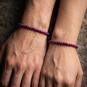 May include: Two matching bracelets featuring small, faceted, deep magenta-coloured beads. Each bracelet consists of a single strand of uniform, round, polished stones that reflect light across their multiple cut surfaces, worn around the wrists.