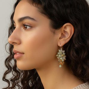 May include: A person wearing cluster earrings. The earrings feature small, round beads in shades of light green and amber, clustered together to resemble grapes. They hang from silver hooks. The earrings are a unique accessory.