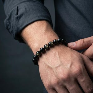 May include: A black beaded bracelet with golden flecks. The bracelet is made of round, polished beads and is worn on a wrist. The beads have a reflective quality, and the bracelet is set against a dark background.