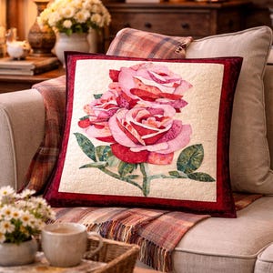 Peut inclure: Un coussin carré décoratif avec un motif floral de roses roses et rouges et de feuilles vertes. Le coussin a un fond crème et une bordure rouge foncé. Il est posé sur un canapé beige avec une couverture à carreaux.