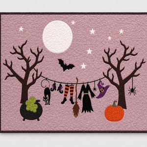May include: A rectangular wall hanging featuring a textured mauve background with a night scene. Two dark trees frame a clothesline holding a black cat, striped stockings, a broom, a dress, and a witch hat. A full moon, stars, a bat, a pumpkin, and a cauldron are present.