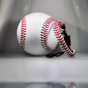 Puede incluir: Una pelota de béisbol blanca con costuras rojas y un cordón de cuero. La pelota está enfocada, con sus costuras rojas y su superficie texturizada claramente visibles. El cordón está envuelto alrededor de la pelota.