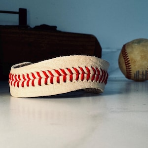 May include: A baseball-themed leather bracelet with red stitching, resembling the seams of a baseball. The bracelet is off-white and sits on a white surface, with a baseball in the background.