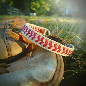 May include: A baseball-themed bracelet with a white band and red stitching, resting on a well-worn baseball glove. The bracelet's design mimics the stitching of a baseball. The glove is a light brown color, suggesting it's been used.