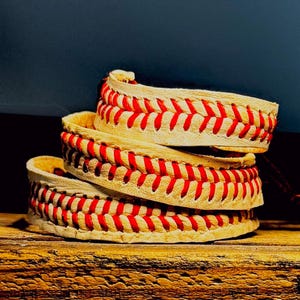 Puede incluir: Tres pulseras apiladas con temática de béisbol. Cada pulsera es de color crema con costuras rojas, que se asemejan a una pelota de béisbol. Las pulseras están apiladas sobre una superficie de madera, con un fondo azul oscuro.