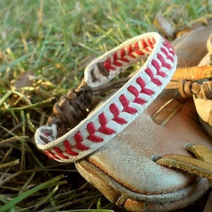 May include: A baseball-themed bracelet with a white band and red stitching, resembling a baseball. The bracelet is set against a backdrop of a baseball glove and green grass, evoking a sporty, outdoor theme.