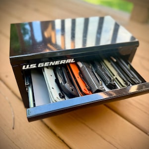 May include: Black metal toolbox with an open drawer revealing various tools. The text "U.S. GENERAL" is visible on the front of the toolbox. The tools include knives and other implements, with orange and black handles.