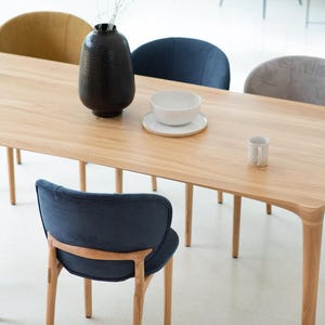 Può includere: Un tavolo da pranzo rettangolare in legno chiaro con angoli arrotondati, circondato da sei sedie con sedute imbottite di vari colori. Un vaso nero, una ciotola bianca e una tazza sono sul tavolo. Le sedie hanno gambe in legno.