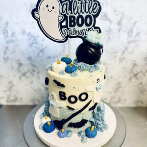 May include: A two-tiered cake decorated with a blue and white frosting. The cake is decorated with ghosts, bats, pumpkins, and a black cauldron. The cake topper says "a little boo is almost here".